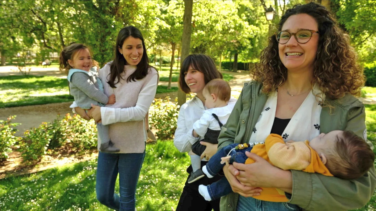 Three mothers holding their babies walk through a leafy and sunny park.