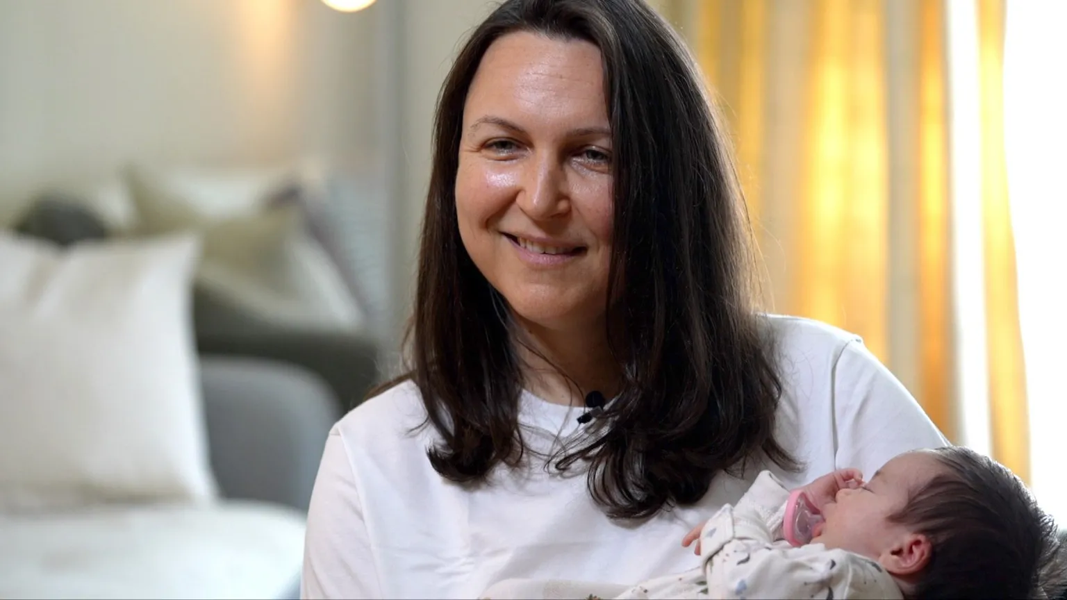 Ana is holding her baby, she is wearing a white top and has long brown hair.