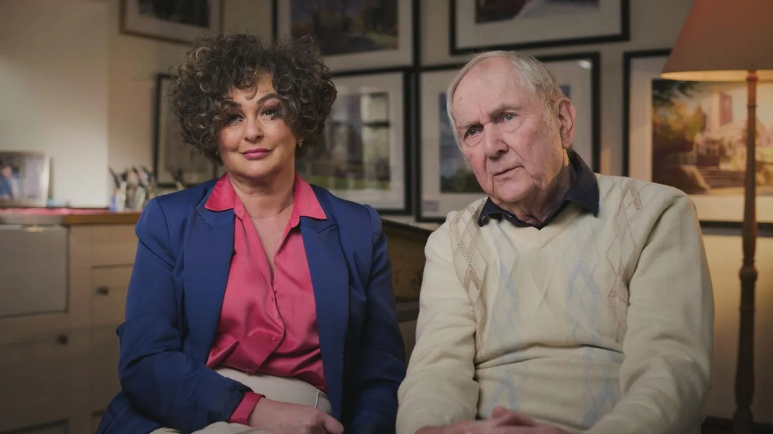 BBC CRIMEWATCH Melanie's father Steve and sister Dominique sitting for a BBC Crimewatch interview. Dominique is wearing a bright pink silk blouse under a dark blue blazer, and has short dark curly hair. Steve is wearing a brown collared shirt underneath a patterned beige jumper. In the background there are lots of framed pictures hung on the wall and a beige floor lamp.