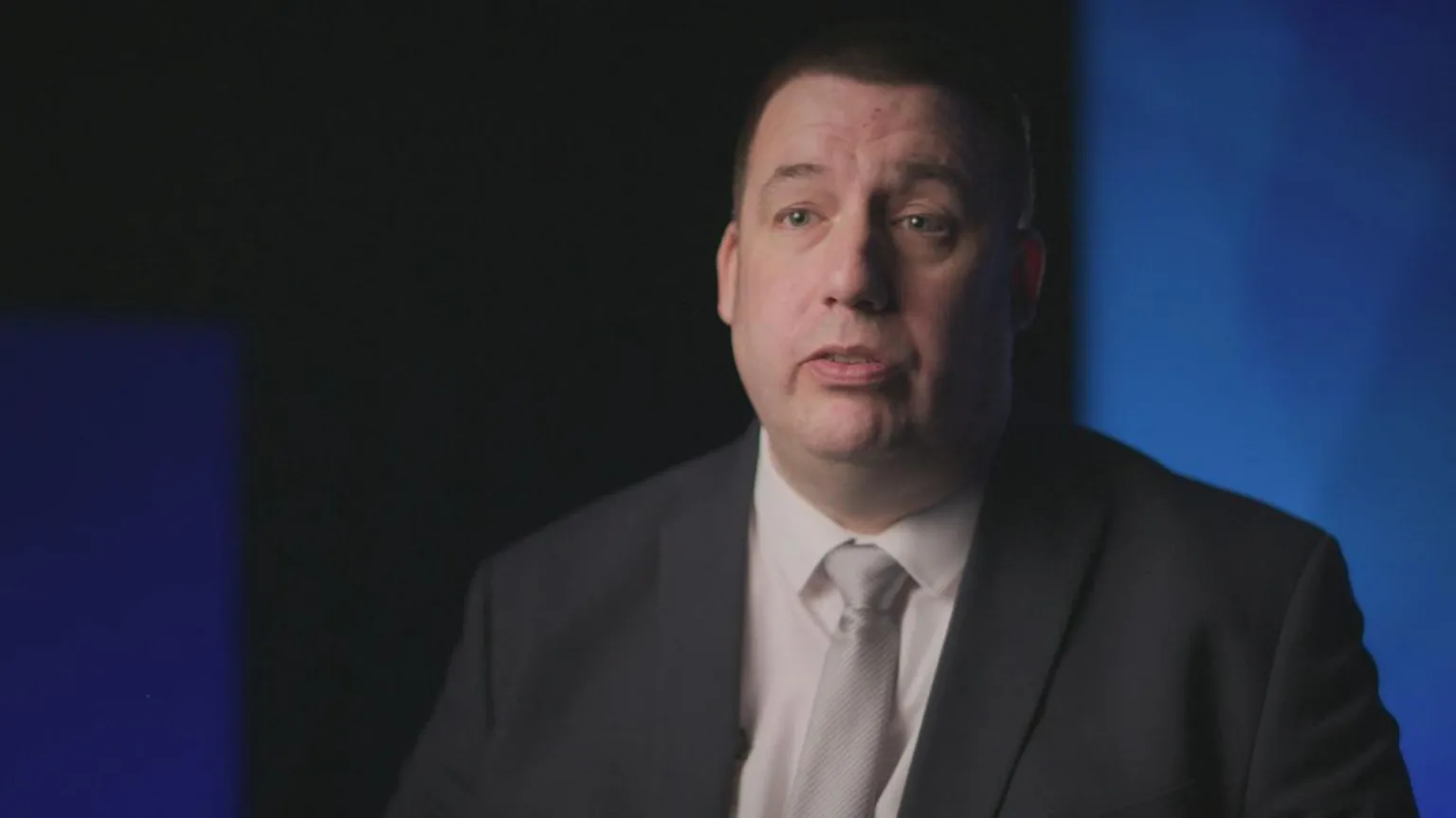 BBC Crimewatch DCI Ben Lavender wearing a white shirt, light grey tie and black suit. He is sitting in front of a blue background, giving an interview for BBC Crimewatch.