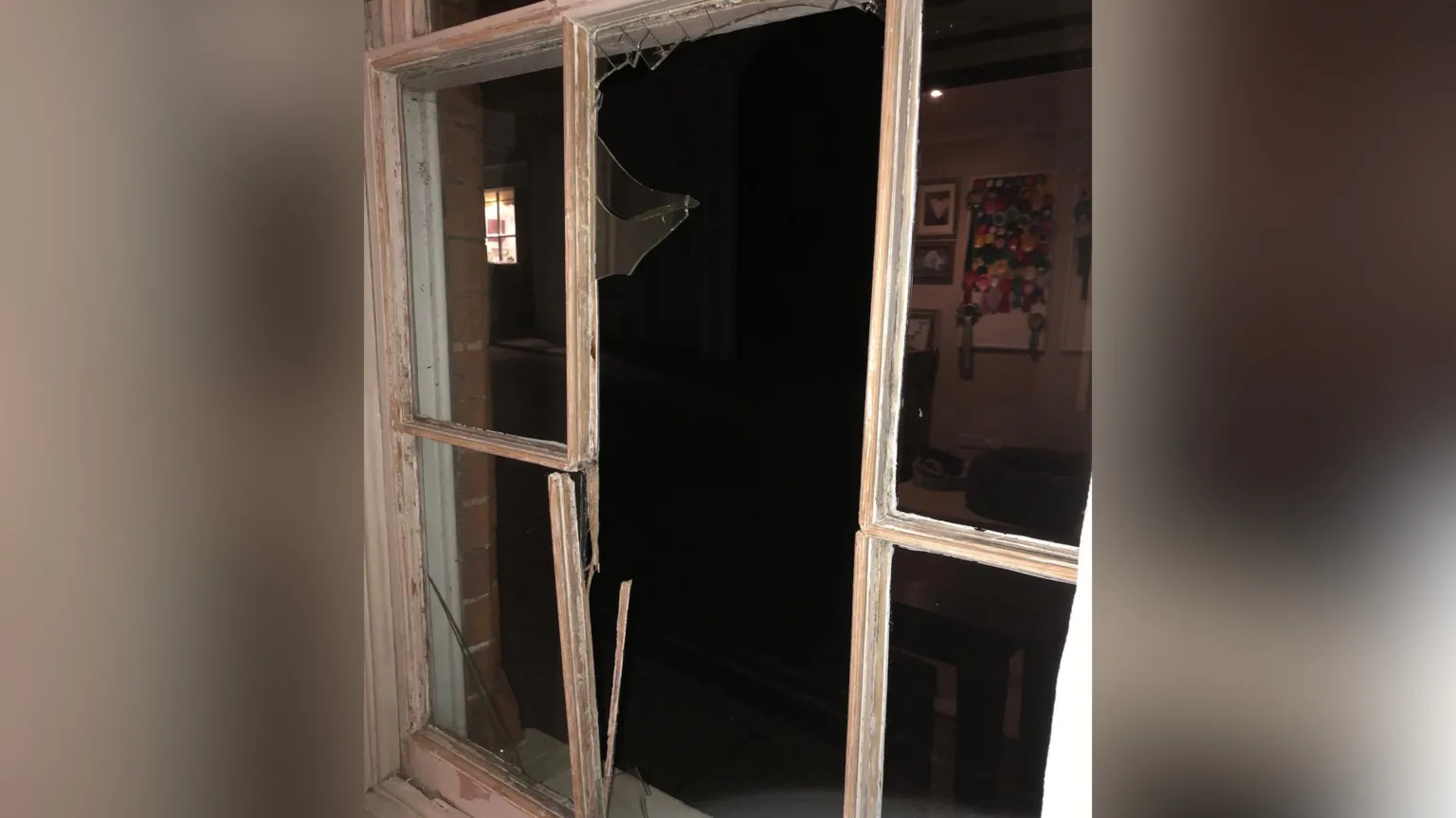 Supplied A large white, wooden window frame with its glass smashed out. The image is taken from inside the home on which the window is attached, showing the outside, which is dark. 
