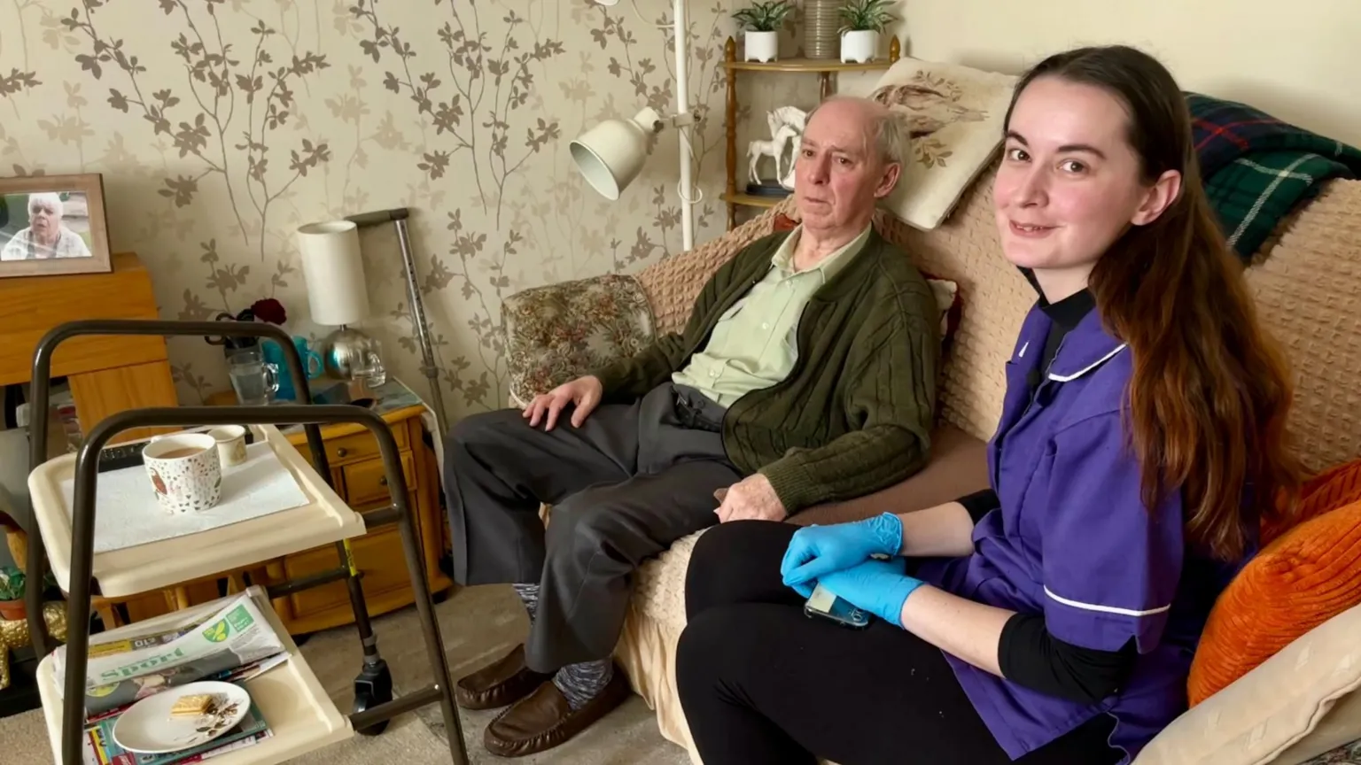 JAMIE NIBLOCK/BBC Mollie Cole-Wilkin sits inside the home of Stephen Mears. Stephen is sitting in a room with leaf-patterned wallpaper. There is a metal framed walker to the left of the picture with an empty plate and cup on a tray. Mollie is wearing a purple carers uniform with white piping and has blue gloves on. Stephen has a dark green cardigan with a light green shirt on and grey trousers, he is wearing brown slippers. There is a picture of his late mother in the background.