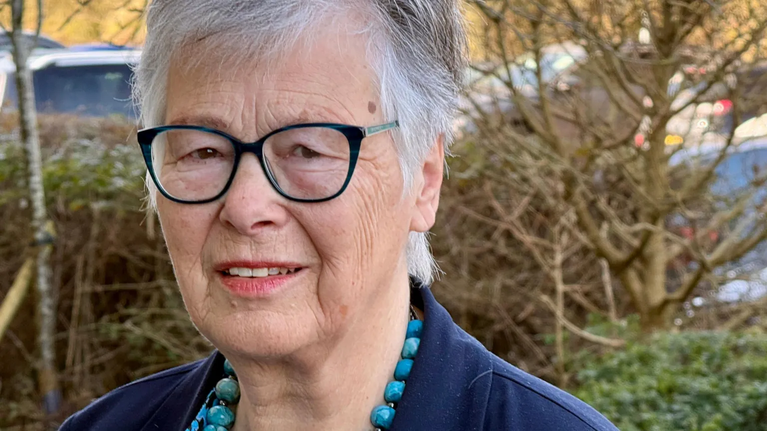 JAMIE NIBLOCK/BBC Janet Beacham stands outside her office in Colchester. She is in front of a hedgerow and a few trees without leaves. There are cars parked behind that. She is wearing a navy blue jacket and has a light blue mottled shirt and matching blue beads around her neck.