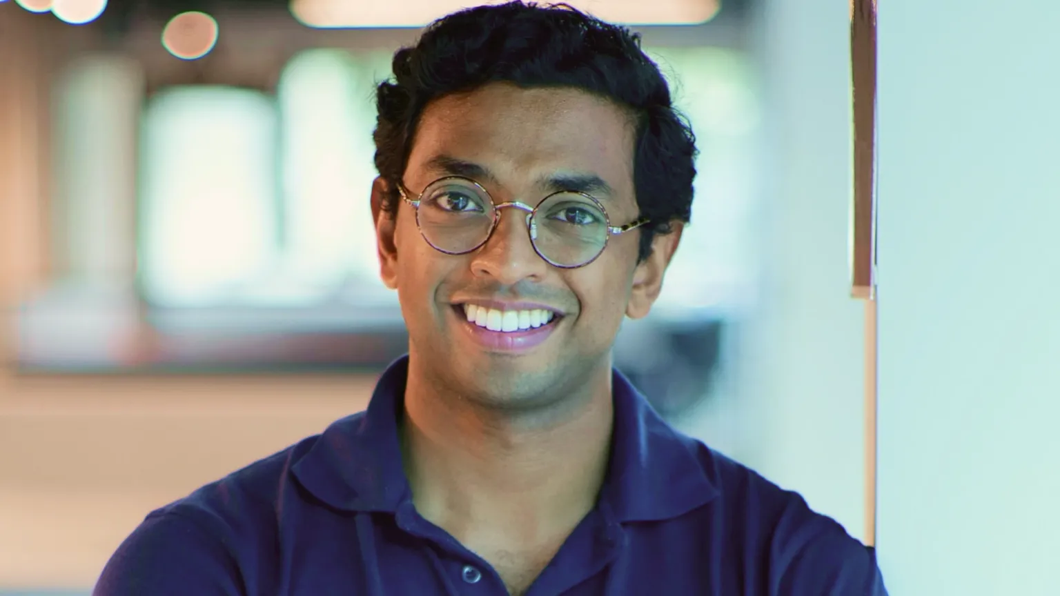 Cera Dr Ben Maruthappu looks at the camera wearing a purple polo shirt. He is smiling and has round metal framed glasses on. He has black hair. The background is a blurred office.
