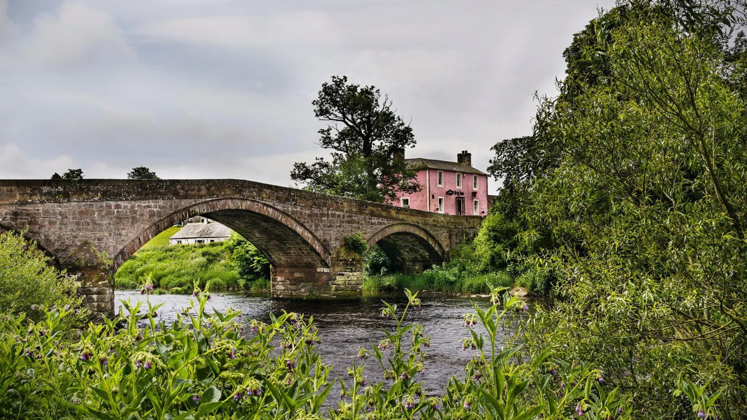 James Johnstone A pink village pub next to a bridge with water flowing underneath it