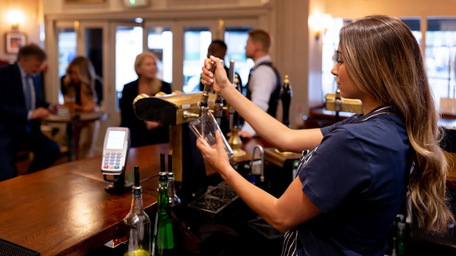  A female member of bar staff pulls a pint of bear in a half-crowded bar. 