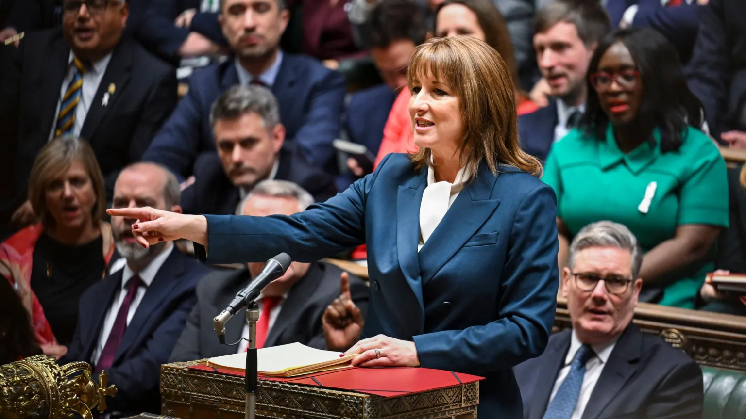 House of Commons/PA Wire Chancellor Rachel Reeves delivers the Budget in the House of Commons on 26 November 2025. She wears a blue suit and is pointing with her right hand, index finger extended. Prime Minister Sir Keir Starmer sits behind her on the front bench, with other Labour MPs visible, listening to Reeves' speech. 
