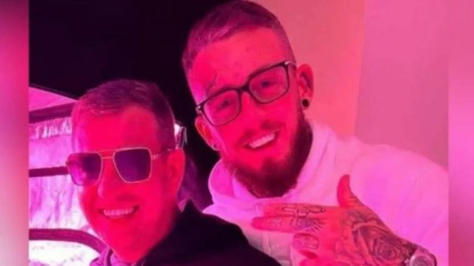 Facebook Two men are smiling at the camera. On the left is John George he is wearing dark glasses and a dark hoodie. On the right is another man wearing glasses and a white hoody. He has his hands up, his hand has tattoos and he is wearing a large wristwatch. He also has a small tattoo above his right eye. 
