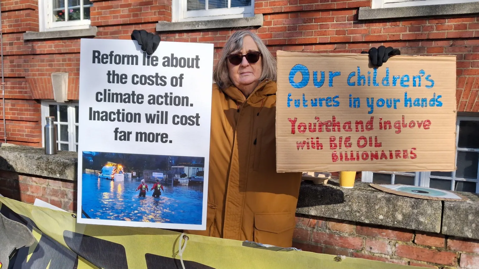 LDRS A woman with shoulder length grey hair and wearing an orange coat. She is wearing black sunglasses and holding up two signs. One says 'Reform lie about the costs of climate action. Inaction will cost far more' and the other says 'our children's futures in your hands. You're hand in glove with big oil billionaires.