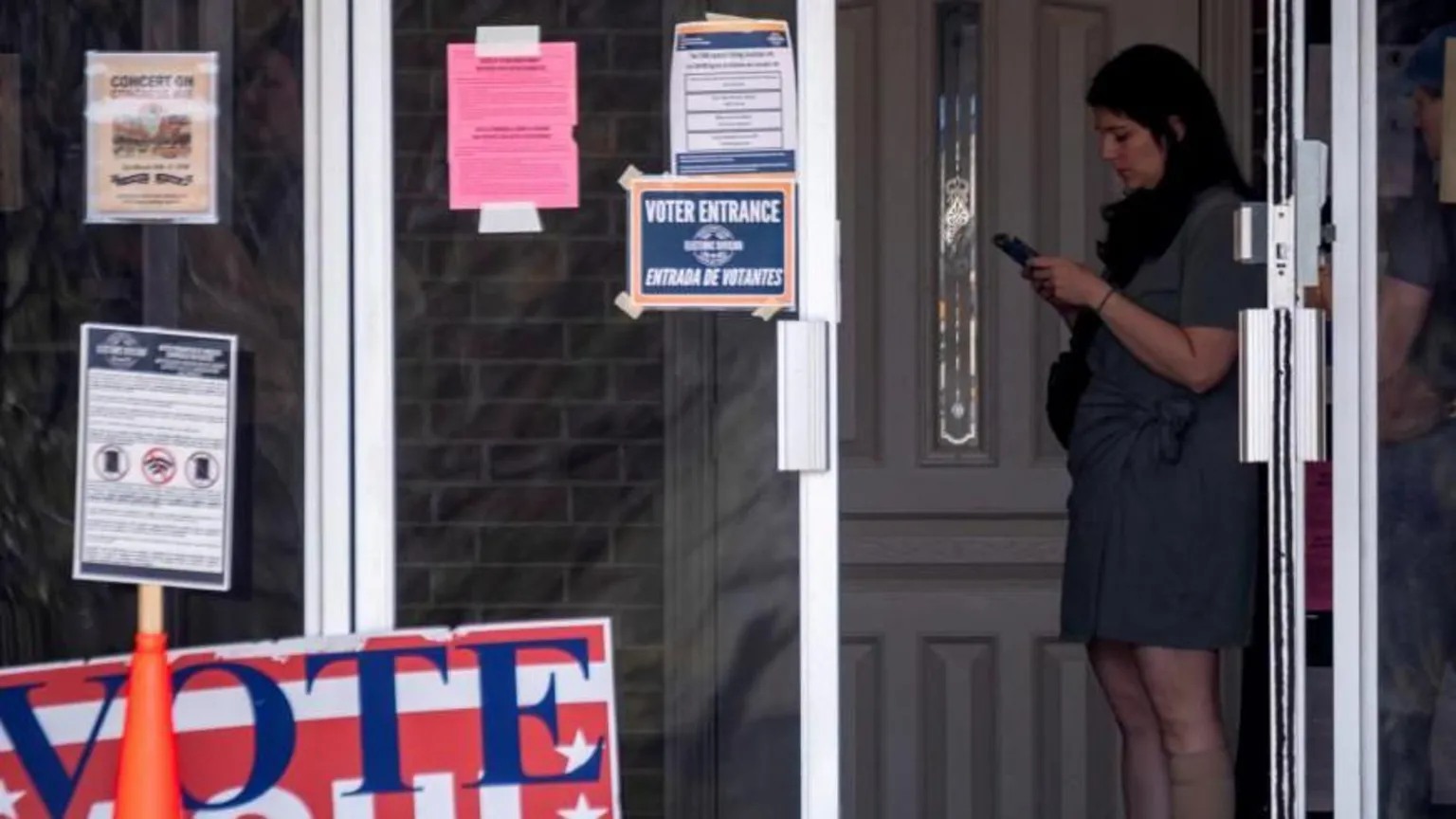  A woman stands looking at here phone at a polling station. A large sign reads 