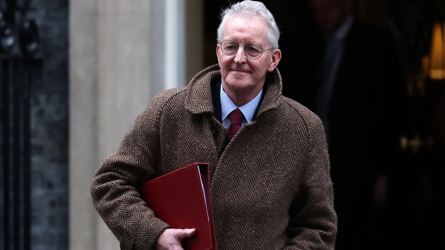 A man with white hair and glasses wearing a brown coat and carrying a red folder