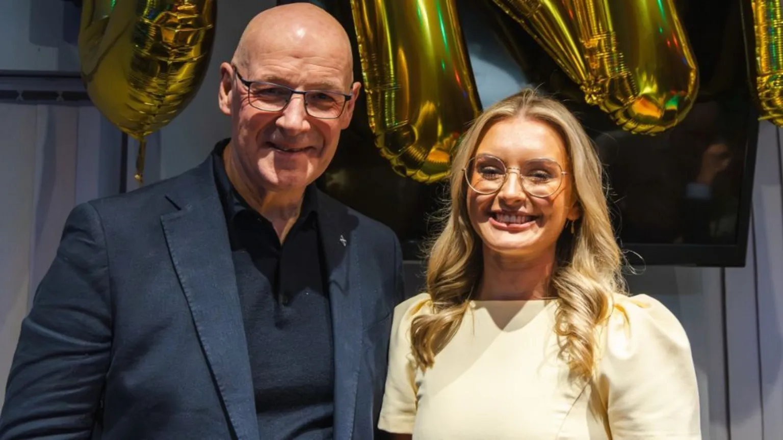 Facebook John Swinney, who is bald with glasses, and Sally Donald, who has long blonde hair and glasses, smile while standing side-by-side. They are posing in front of gold balloons. 