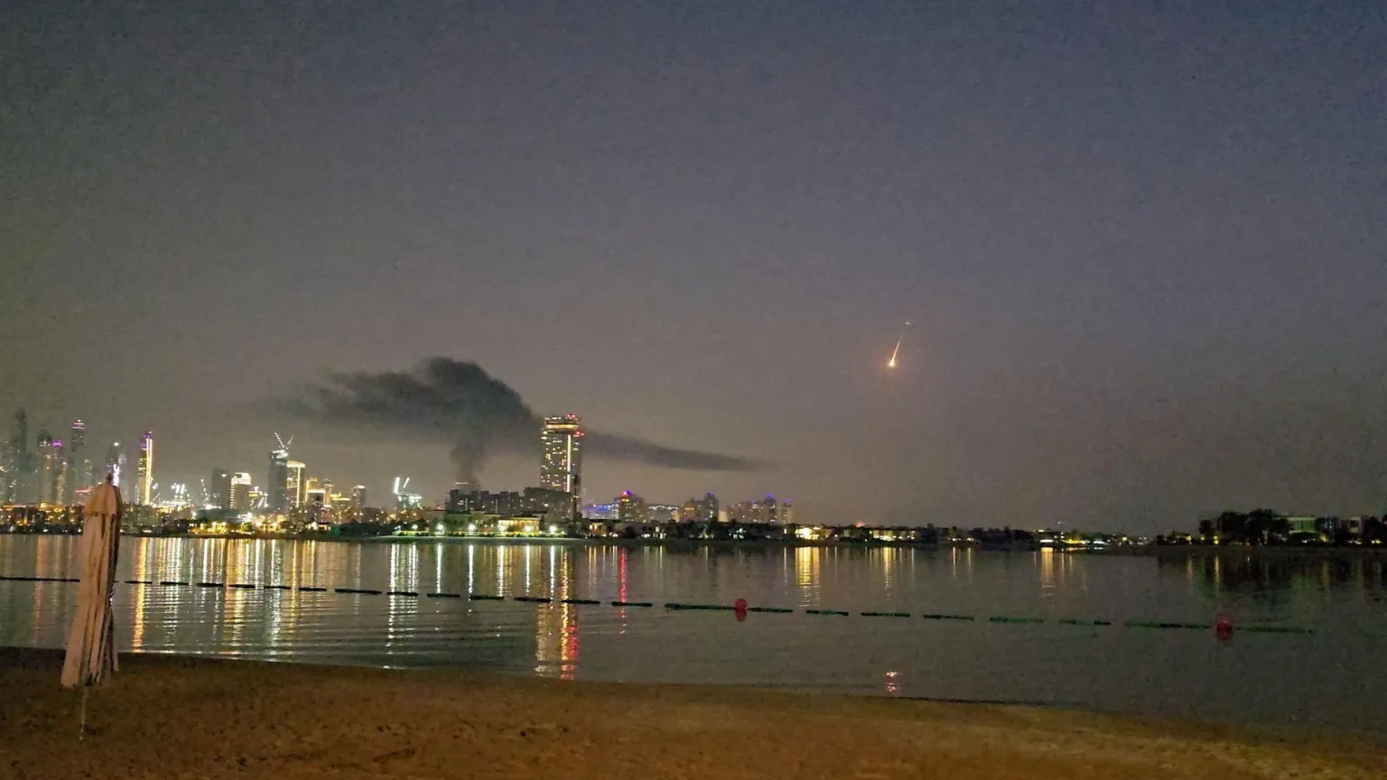  A shot of Dubai's skyline with smoke rising from a high-rise building - the hotel Carson was staying in. It is nighttime and a body of water is reflecting the city lights. The streak of a missile can be seen in the distance.