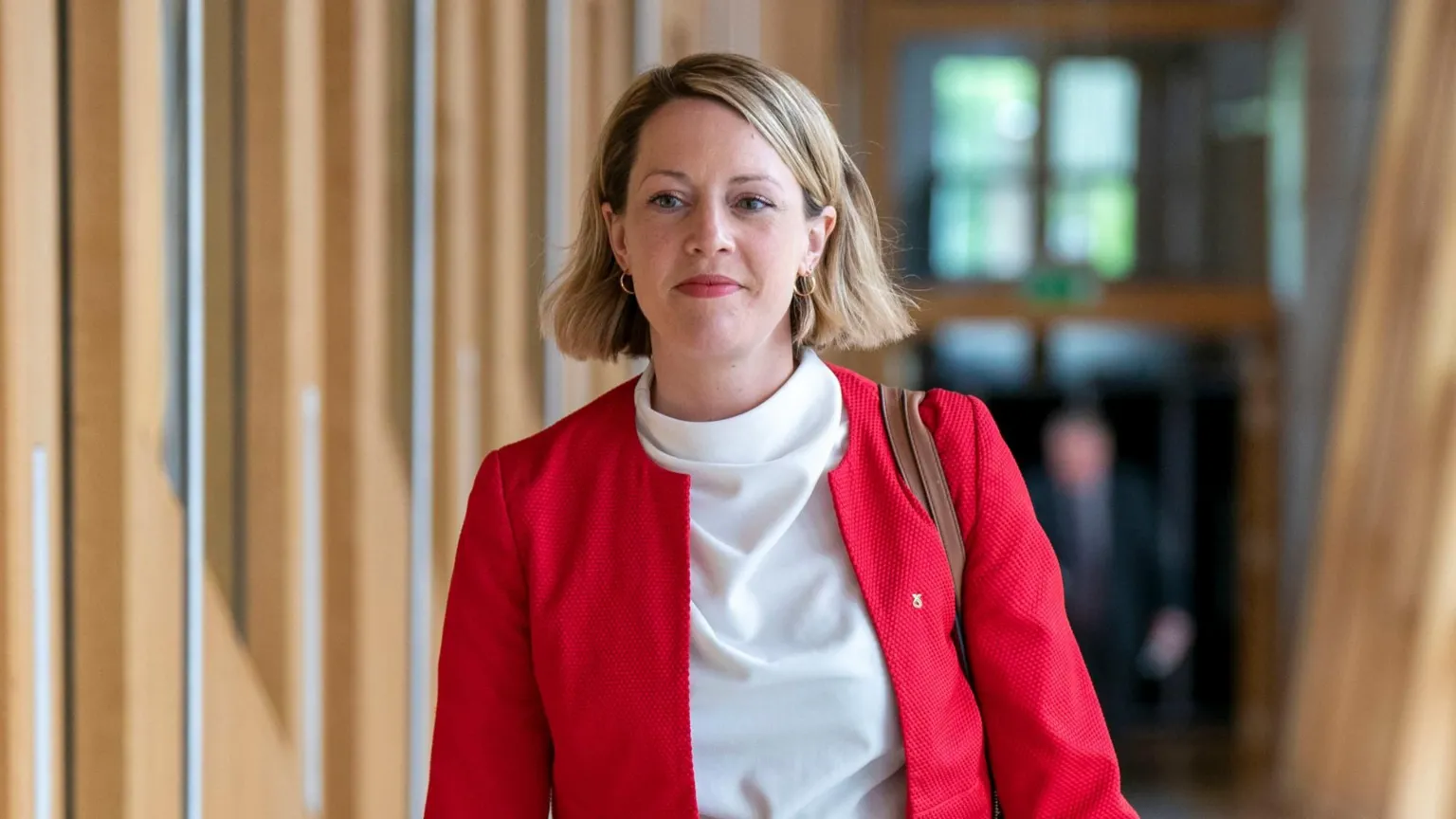 Jenny Gilruth, who has shoulder-length, blonde hair, is walking down a corridor at Holyrood. She is wearing a white top and red jacket.