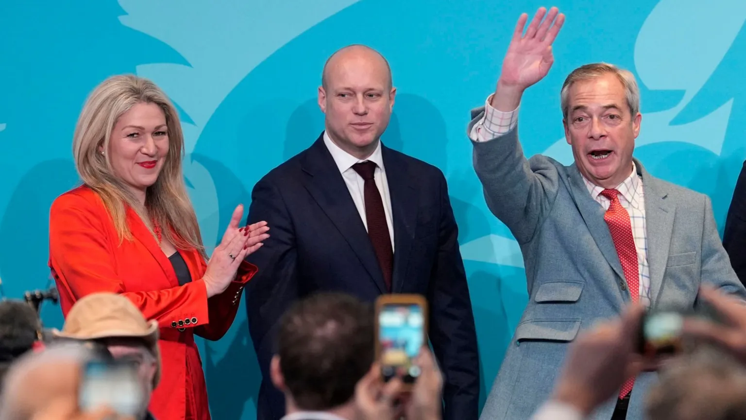  A woman with blonde hair in an orange jacket, Laura Ann Jones, a bald man in a navy suit, Dan Thomas, and a man with grey hair and a light blue suit, Nigel Farage, stand on stage at an event.