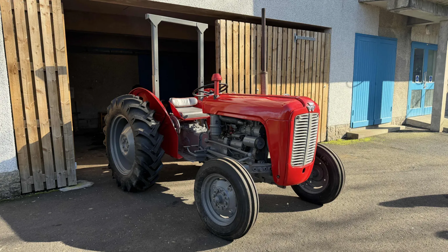 The fully refurbished tractor has been pulled out the garage. The sun shines down on the glossy red paint. the paint is so shiny that you can see reflections in it.