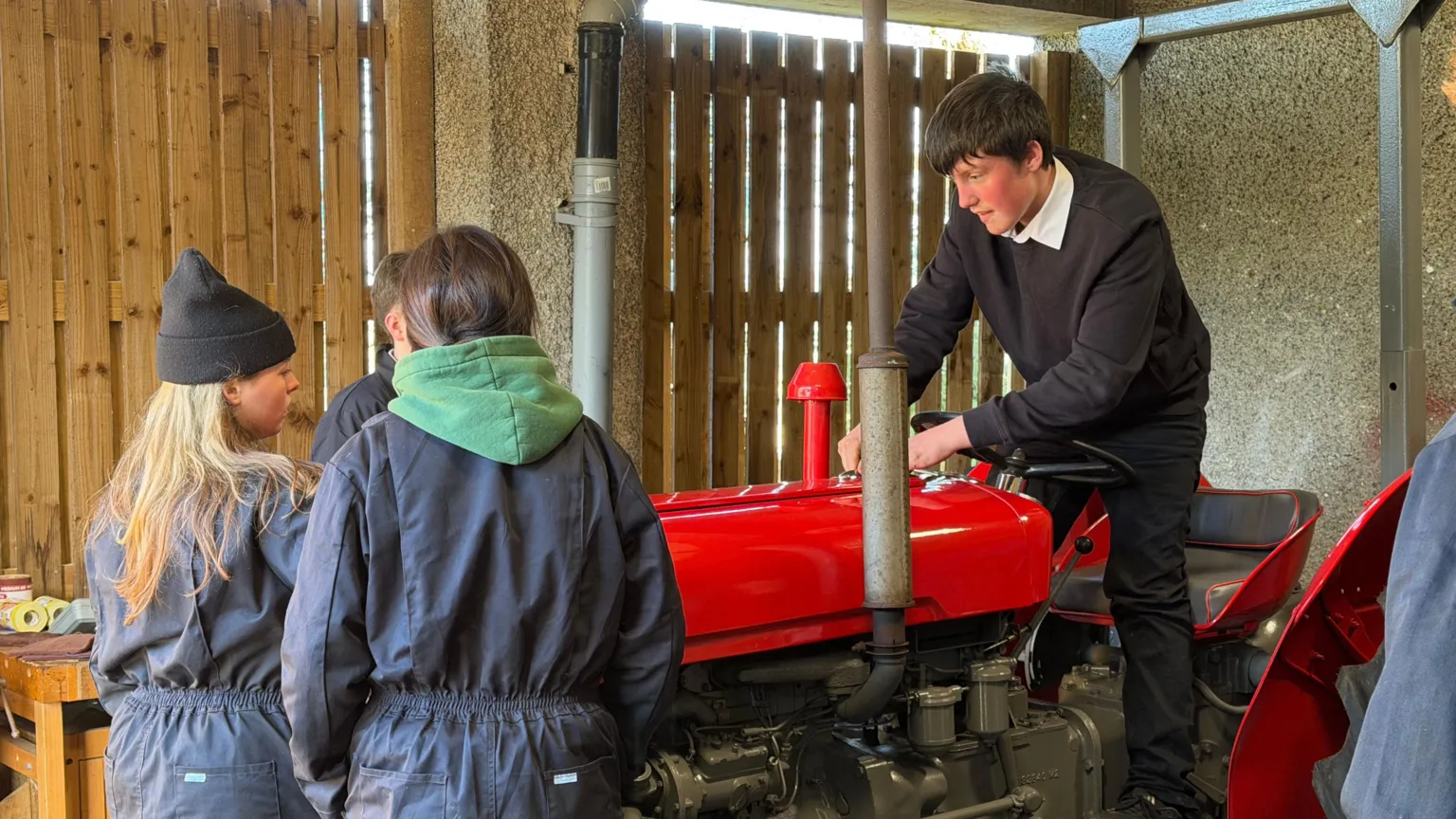 A boy in school uniform is standing up at the seat of the tractor, attempting to turn it on. Three students in boiler suits are standing at the front of the tractor fiddling with something.