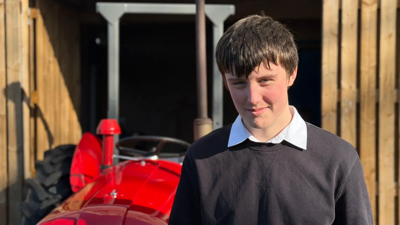 A boy in a school jumper and shirt stands in front of the red tractor. He has straight dark hair and rosy red cheeks. He is squinting as there is a low sun in his eyes.