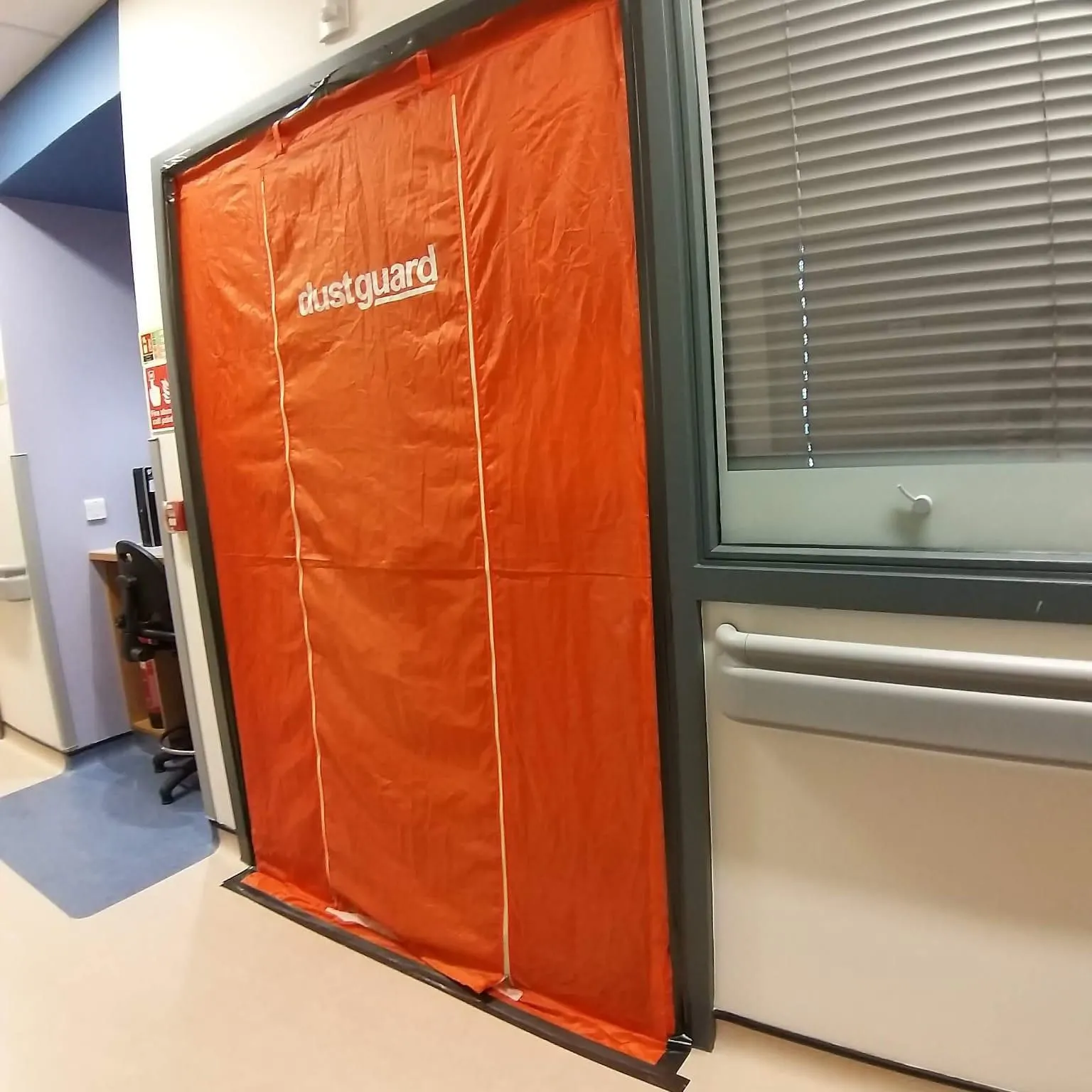 Mark Bissett An orange dust‑containment barrier labeled “dustguard” covers a doorway in a hospital or clinical corridor. The barrier is framed by a grey doorframe, with medical‑style flooring, wall rails, and a window with closed blinds beside it. Part of the hallway and adjacent rooms are visible.