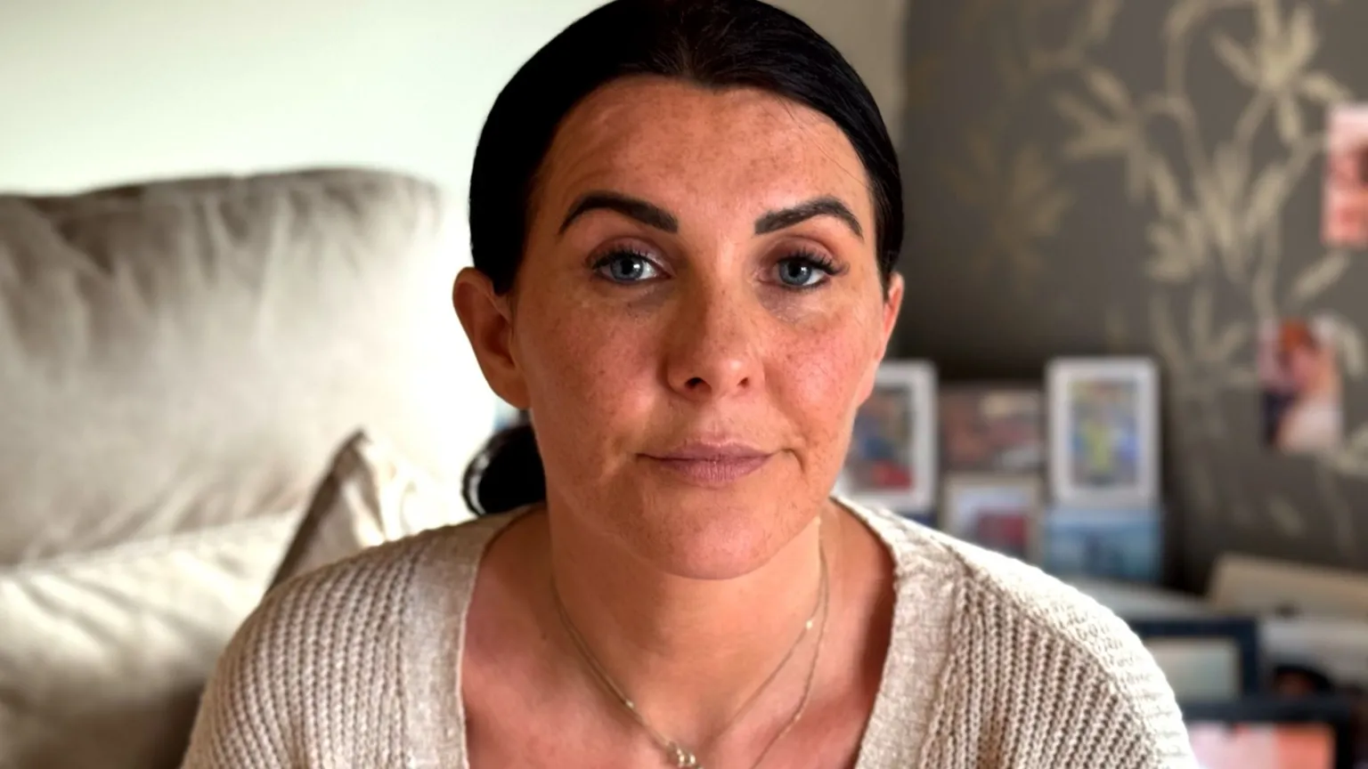 A woman looks into the camera with an expressionless face. Her dark hair is tied back. She is wearing a cream cardigan and thin gold necklace.