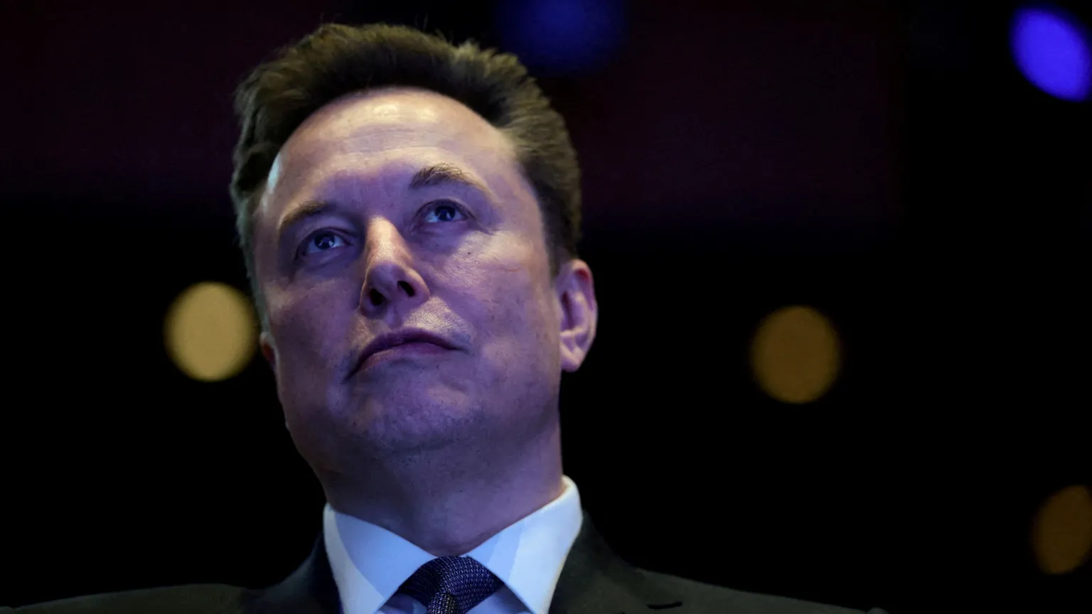 Elon Musk in a black suit and grey neck tie staring out with a bemused look on his face.