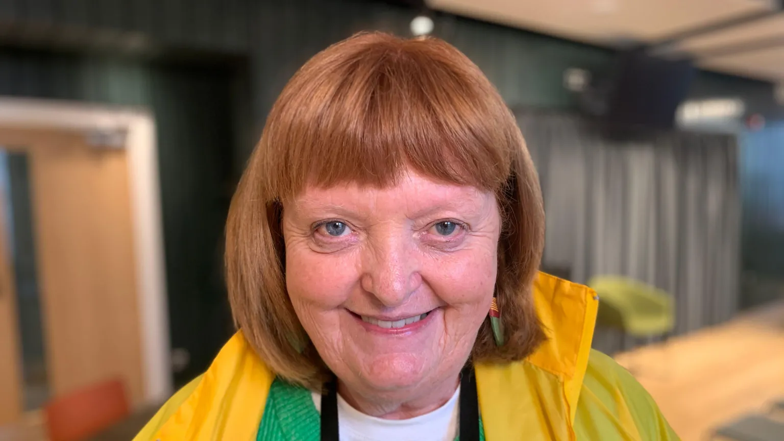 A woman with auburn coloured hair is smiling at the camera. She is wearing a green cardigan and a yellow coloured jacket. 