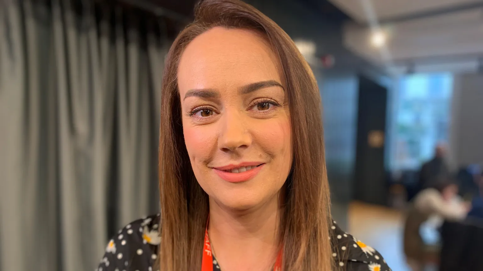 A woman with long brown hair is smiling at the camera. She is wearing a flowery patterned top and is standing in front of a curtain.