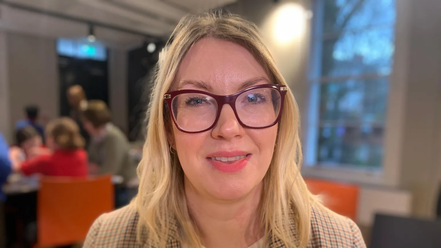 A woman with shoulder length blonde hair and brown framed glasses is looking at the camera. She is wearing a checked blazer with a white top underneath. there are children at desks in the background.