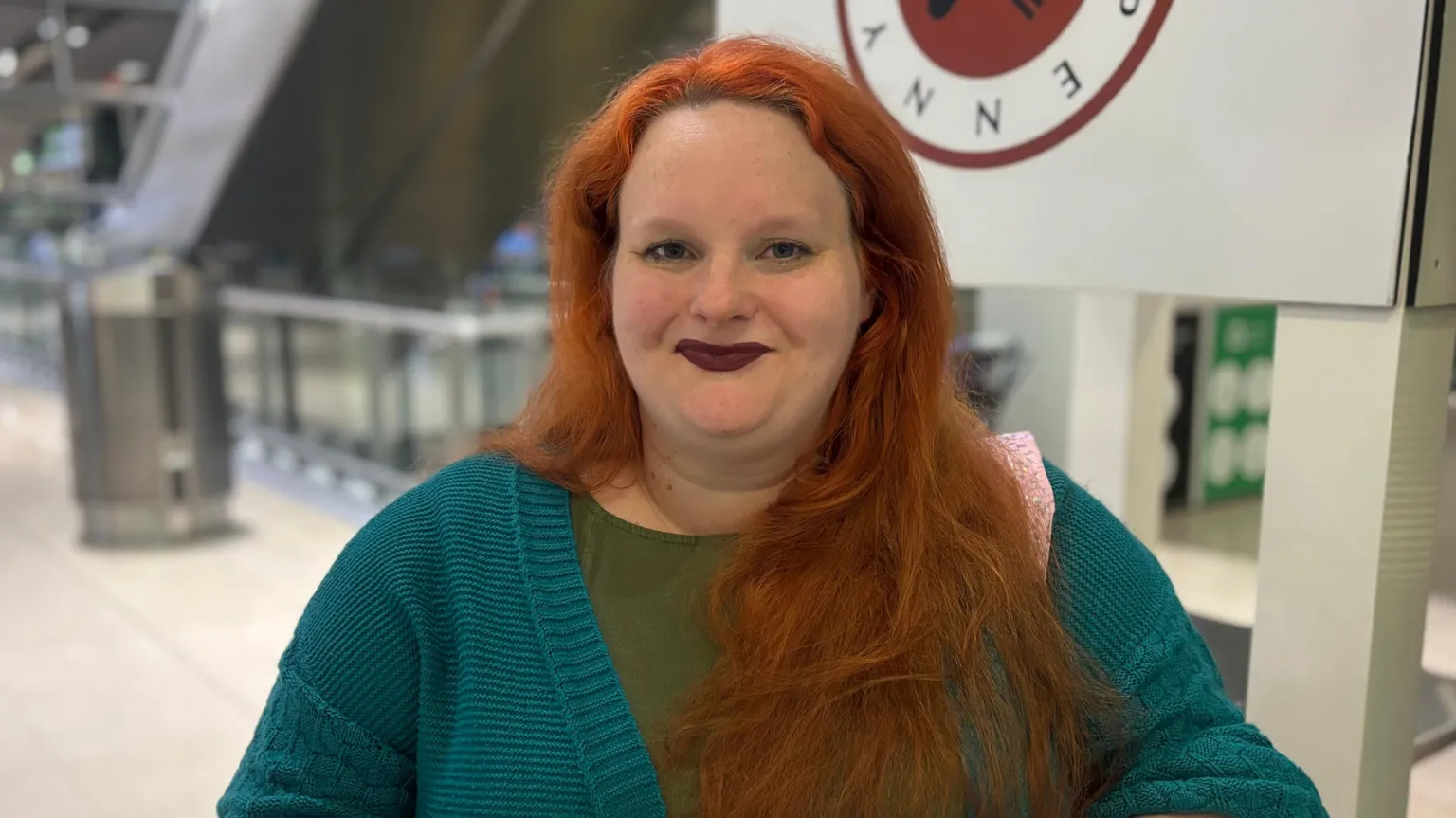 Elizabeth has long ginger hair and dark lipstick. She has a turquoise cardigan and a green top.