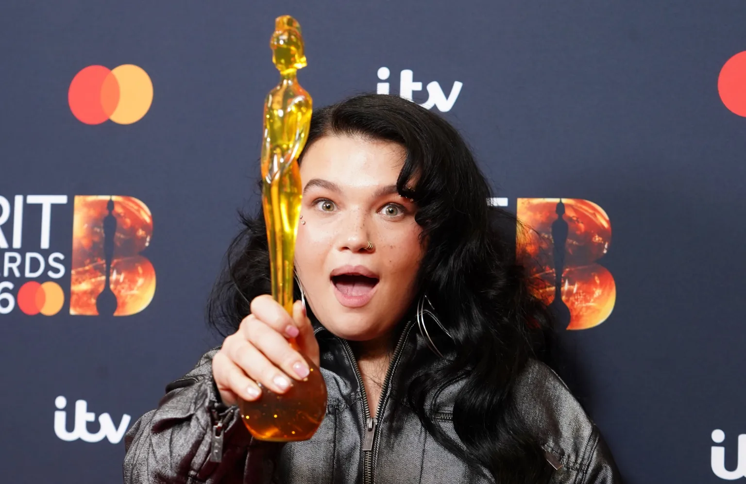  Lola Young holding up the gold Bris statuette in front of her with a shocked expression on her face