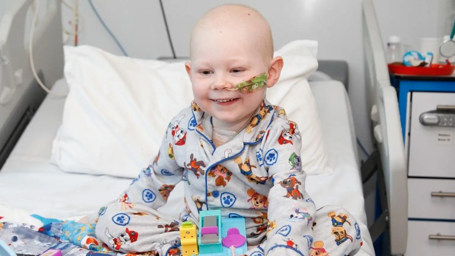 Sarah Kent Vinnie, 4, is sitting on a hospital bed. He has no hair and has a tube going into his nose. Vinnie is smiling and wearing grey and blue pajamas with dogs on them. They are the dogs from a children's TV programme called Paw Patrol. He has a few toys in front of him on the bed.