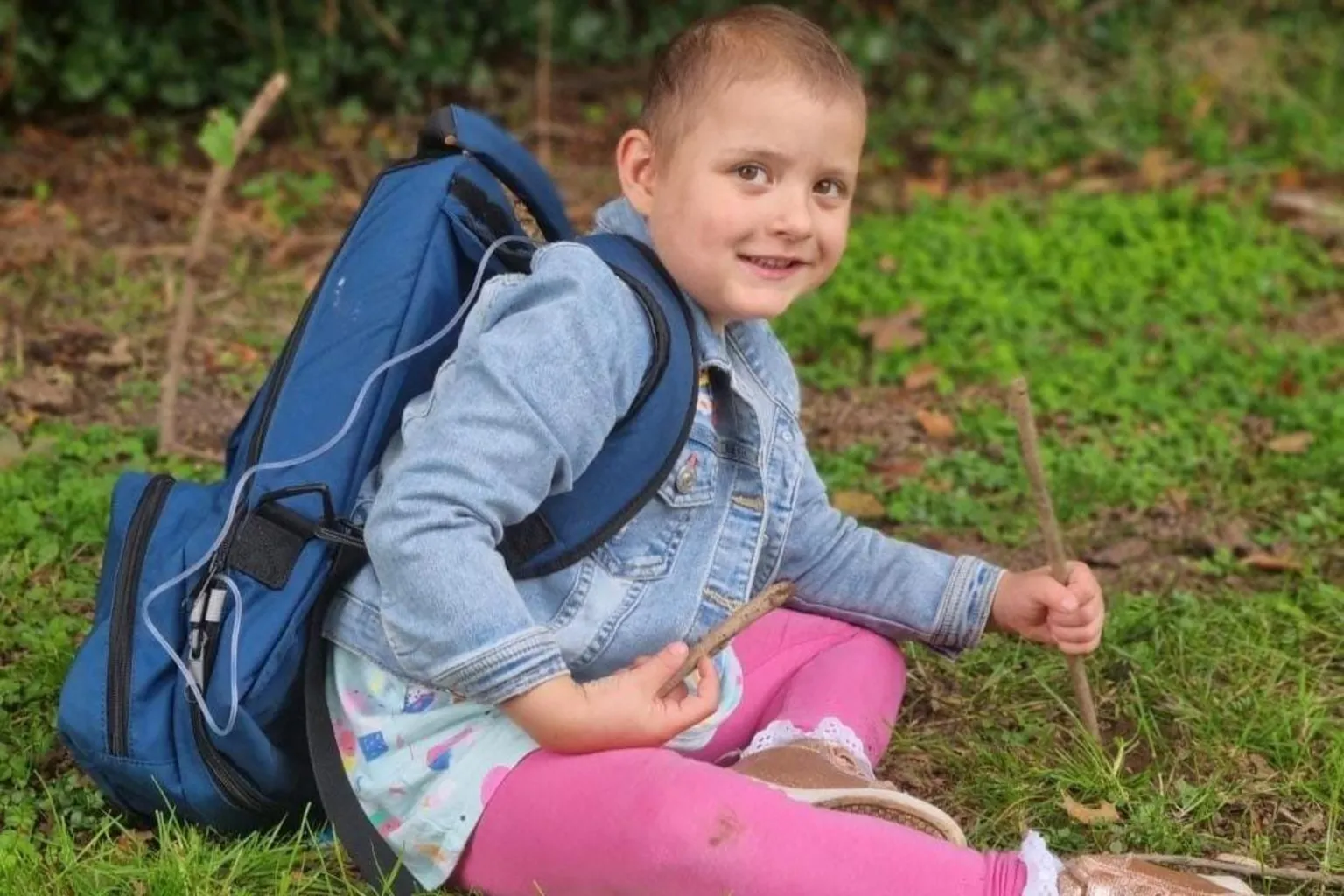 Jess John Lacie, 4, is sitting on the grass and smiling at the camera. She is wearing pink leggings and a denim jacket and holding a stick in each hand. She has a large blue medical backpack on with a pipe going into a port. 