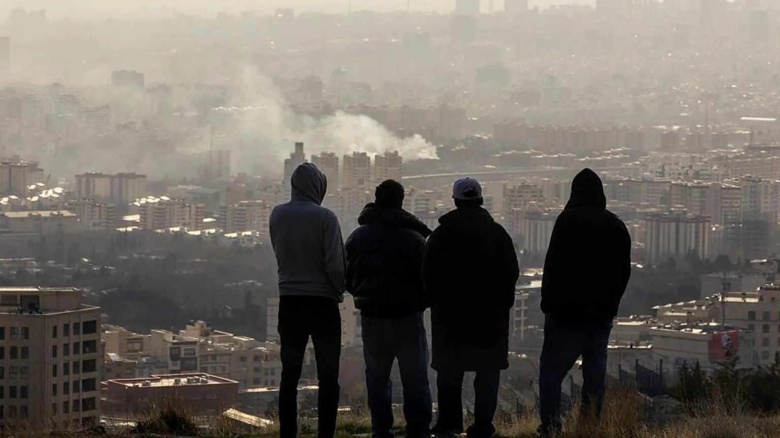  Men watch from a hillside as a plume of smoke rises after an explosion in Tehran, Iran. Beneath them lies the city.