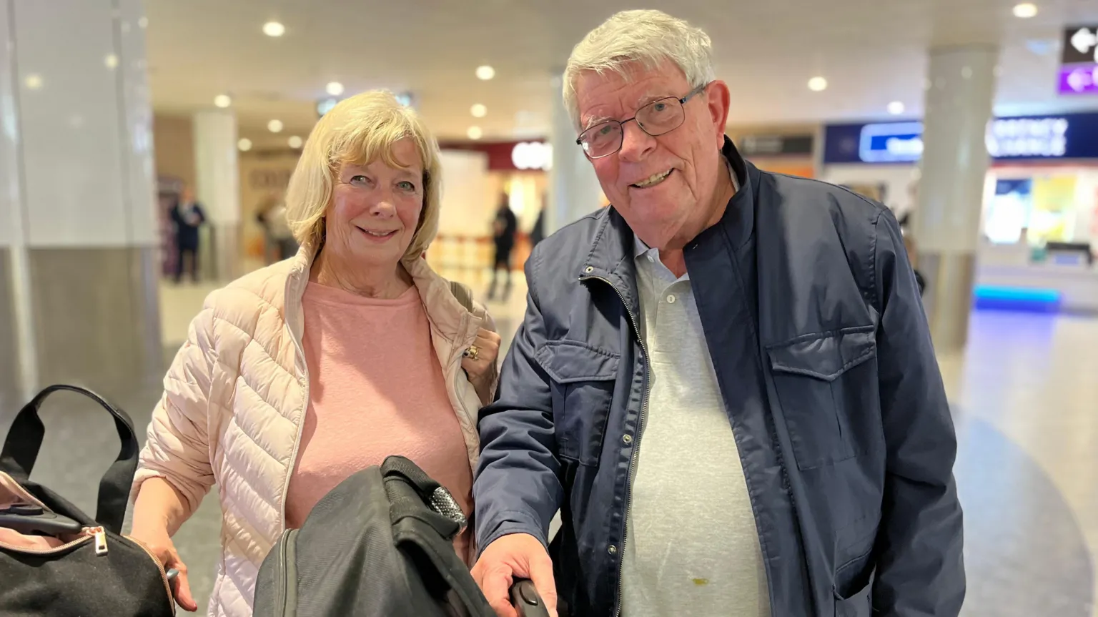 A blonde haired woman in a light jacket and pink top, standing next to a grey haired man wearing a blue jacket in an airport terminal