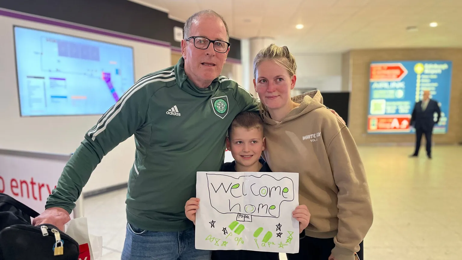 A bespectacled man in a green Celtic top, with his arm around a young boy holding a welcome home sign, and a young woman in a light brown top