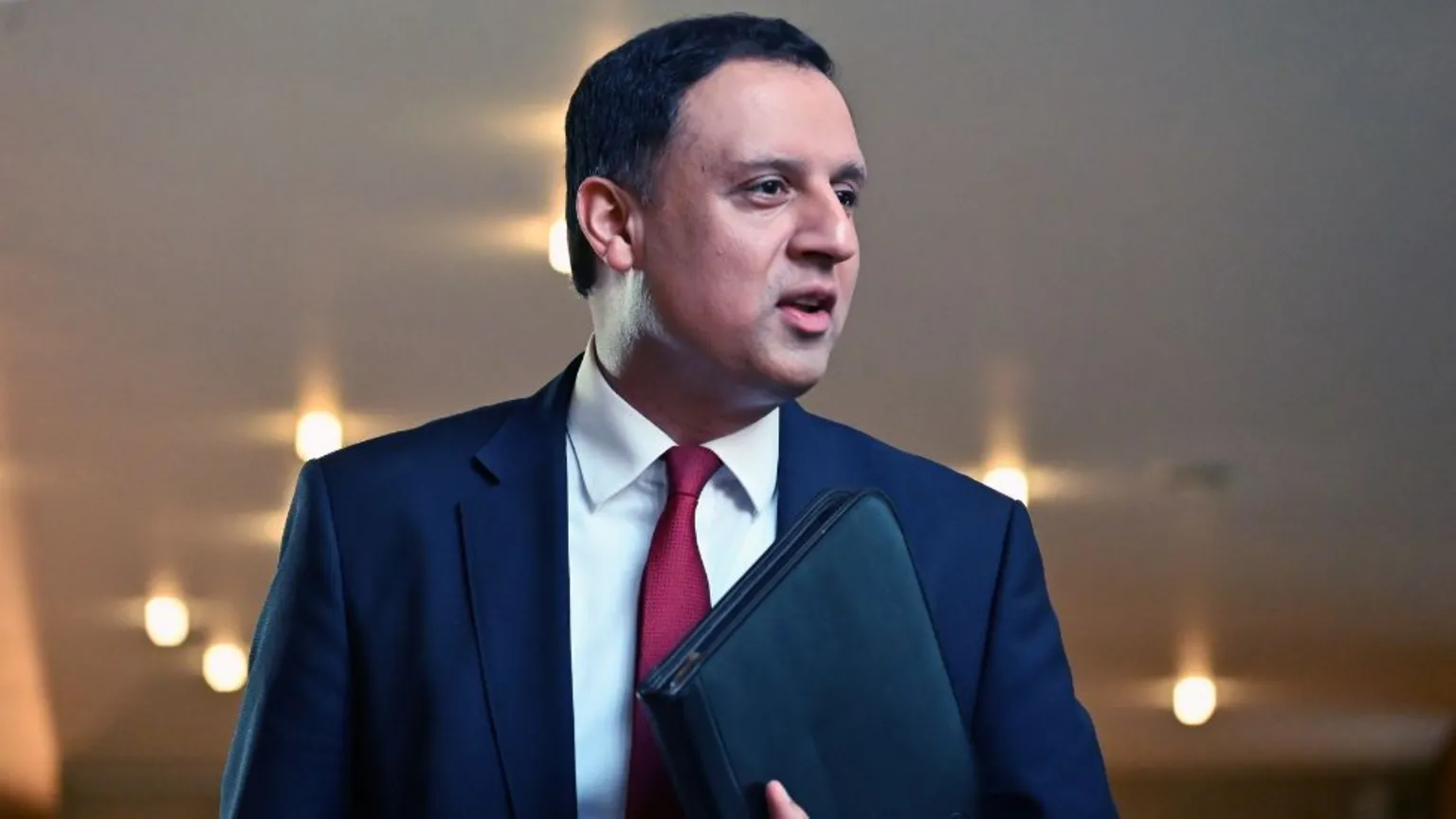  Anas Sarwar, who has short, black hair, looks to his left while carrying a black folder under his arm. He is wearing a navy suit and white shirt with a red tie. 