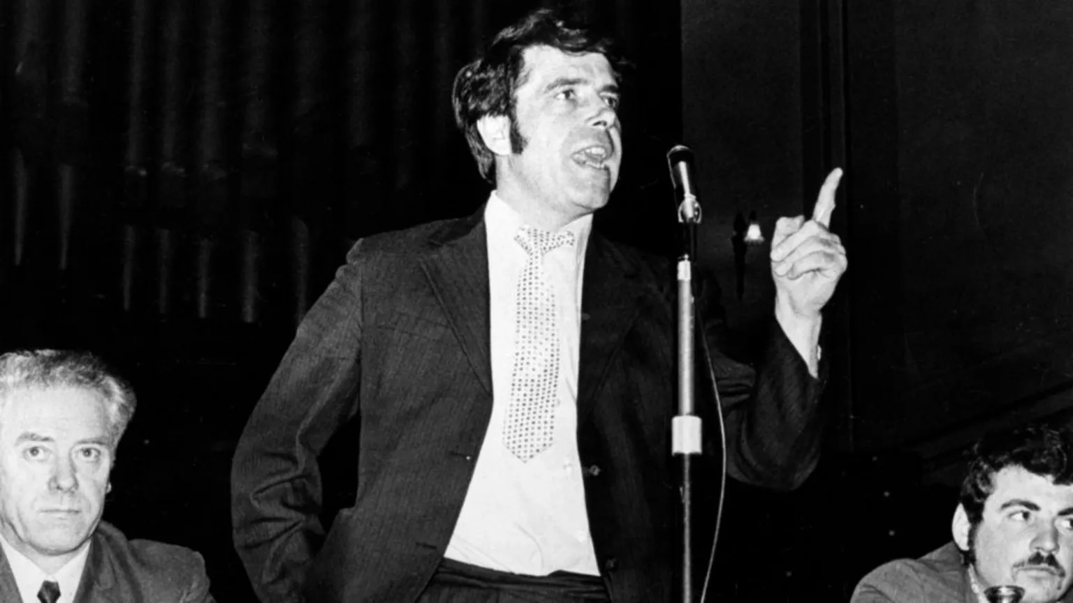  A black-and-white photo of a Jimmy Reid, who has dark hair and side burns, speaking into a microphone. He is wearing a dark suit with a white shirt and patterned tie. He has his left hand raised in front of him, with his index finger pointing upwards. 