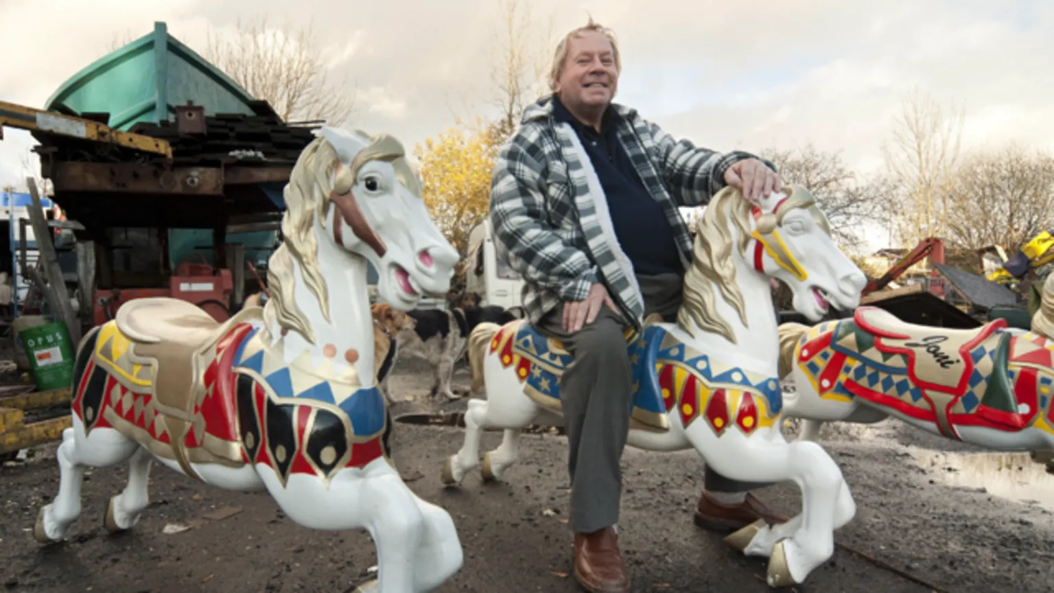 Tom Manley Jimmy Stringfellow in a thick, checked shirt and grey trousers, sits astride a carousel horse with several more on either side of him, within his yard. 
