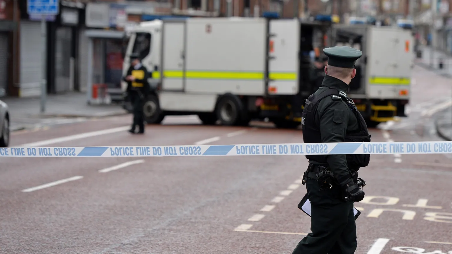  A police cordon has been placed along a street in Belfast city centre. A male police officer in full uniform is standing beside the cordon but looking away from the camera. A female police officer is standing beside a white van which is concealed behind the cordon. 
