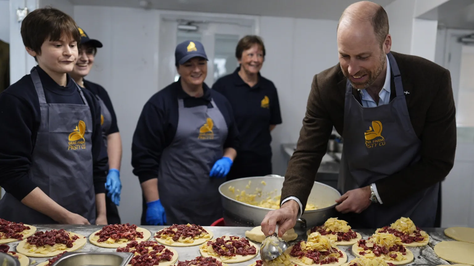  Prince William is holding a pasty. He is wearing a brown jacket and a blue apron.