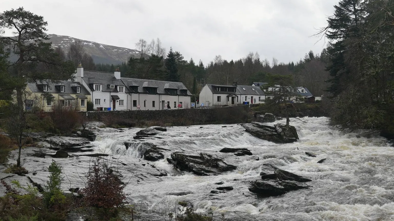 A river flows through jagged black rocks with white foam on the surface. Houses line up by the river wall on the left and trees overhang the water on the right.