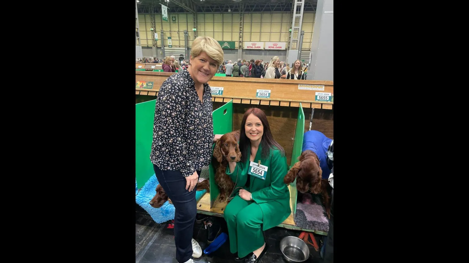 Laura Crombie Two women at Crufts. The one on the left has blonde hair and is wearing a floral shirt and jeans and is smiling at the camera. The second woman is sat down next to a dog in a booth, wearing a green suit.