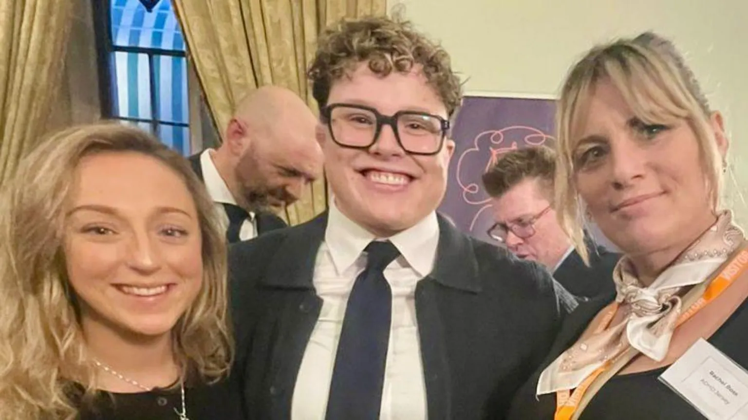 Rachel Boss Charlie, Hannah and Rachel are standing close together at an indoor event, dressed in smart or semi-formal clothing. They are positioned in front of tall curtains and a decorative window, with a banner partially visible behind them. Rachel wears an orange visitor lanyard, and Charlie has her arm around Hannah in the centre.