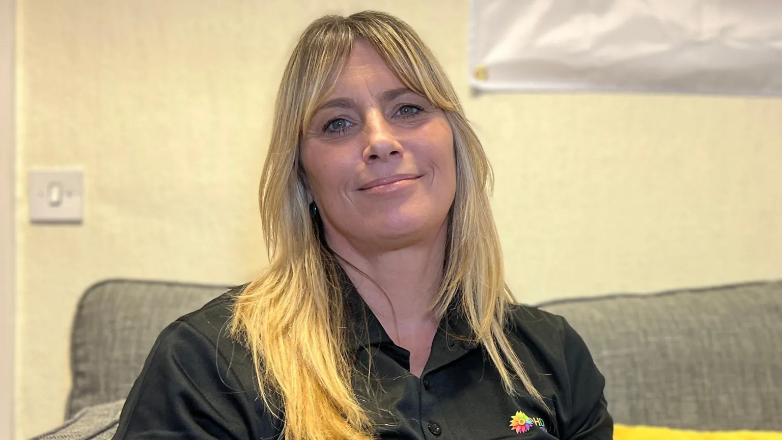 Rachel is sitting on a sofa in a casual indoor setting, wearing a black polo shirt with a small multicoloured logo. A large yellow cushion is placed beside them, and a blank whiteboard or poster hangs on the wall in the background.