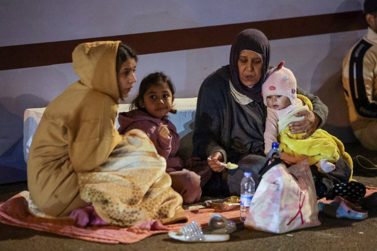 Two women wearing winter clothes sit on a pink blanket, one woman is holding a baby bundled in pink and another is cuddling with a little girl in pink. 