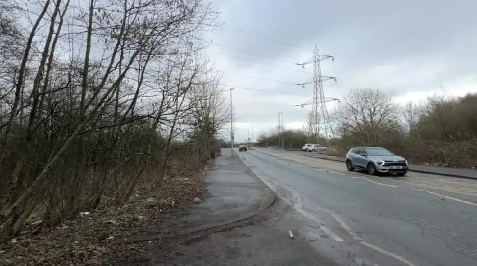 A general view of an isolated stretch of road with three cars visible, and undergrowth and a pathway to the left 