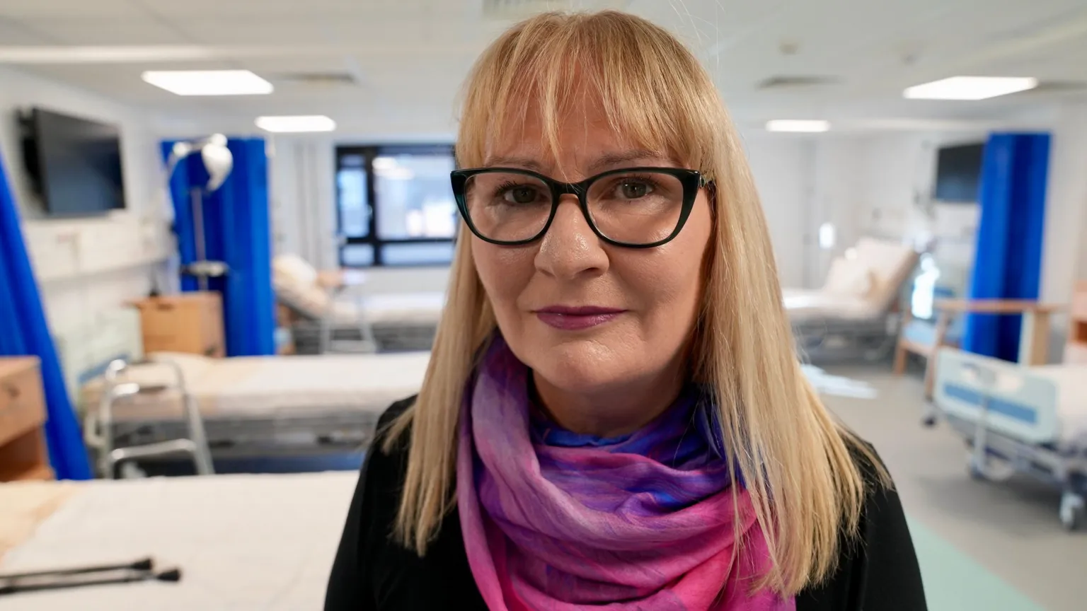 Lesley is wearing glasses, a pink scarf and black top and standing in a practice hospital ward, facing the camera.