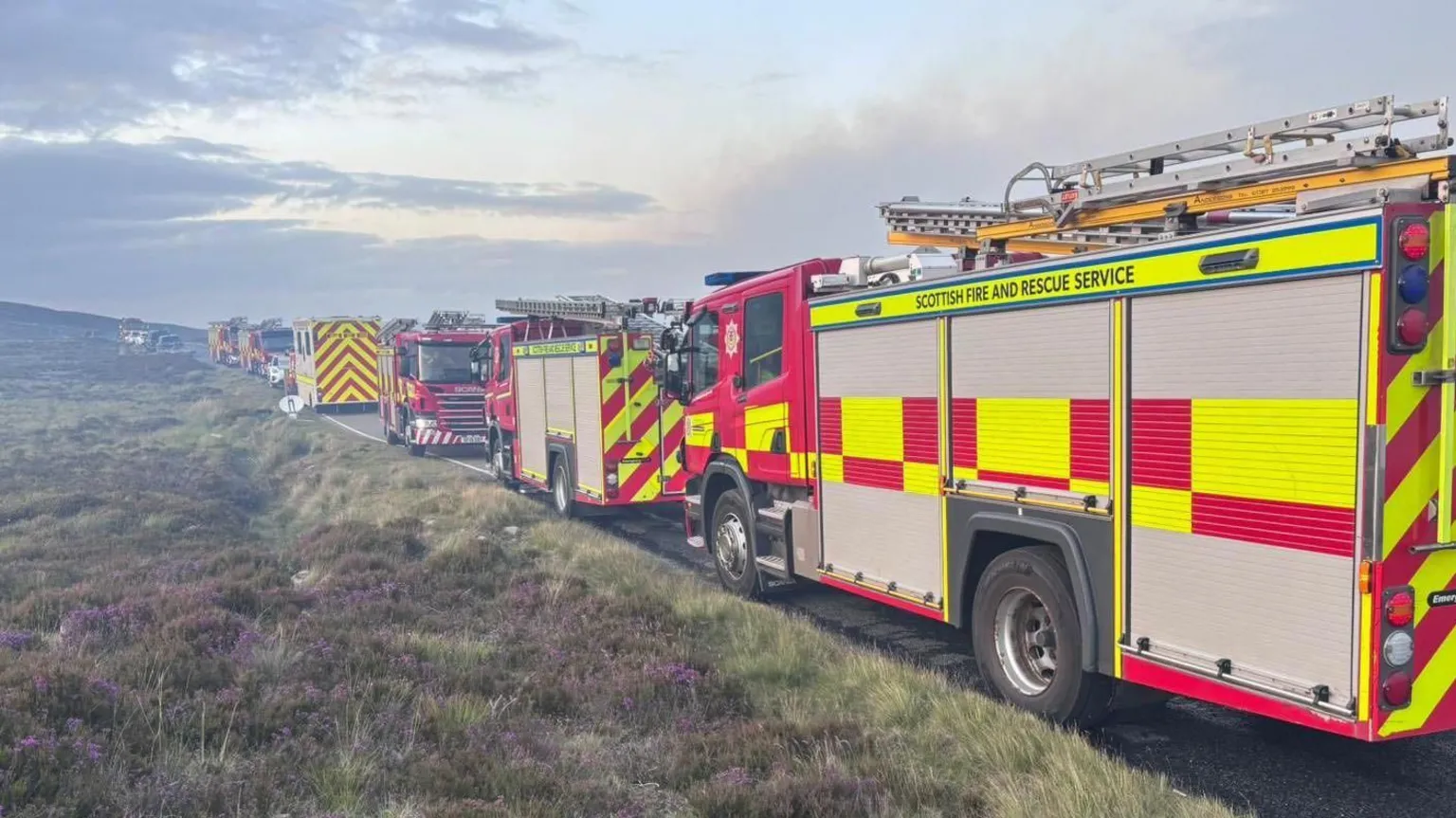 SFRS A row of fire appliances parked by the side of a moorland road. The vehicles are brightly painted yellow and red. Grey smoke drifts overhead. 