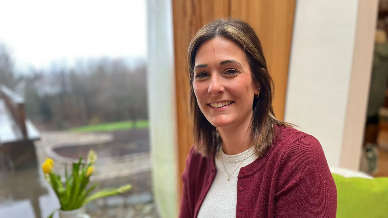 Head and shoulders image of Kirsty Speers. She has shoulder-length, brown hair and is smiling at the camera. She is wearing a white top with a burgundy cardigan. Beside her is a vase containing yellow flowers. 