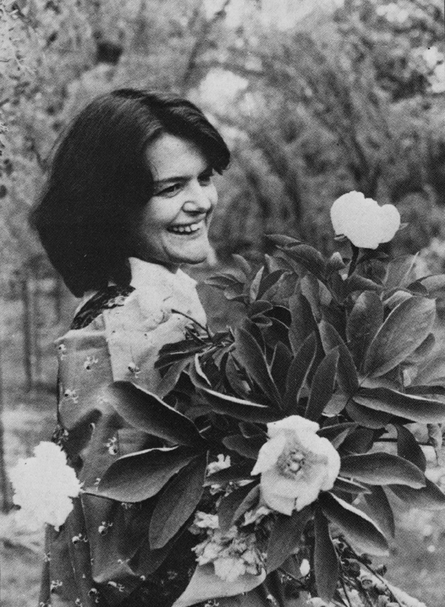 Maggie's Centres Black-and-white image of Maggie Keswick Jencks. She has dark hair and is smiling at something off camera. In front of her is a flowering plant. 